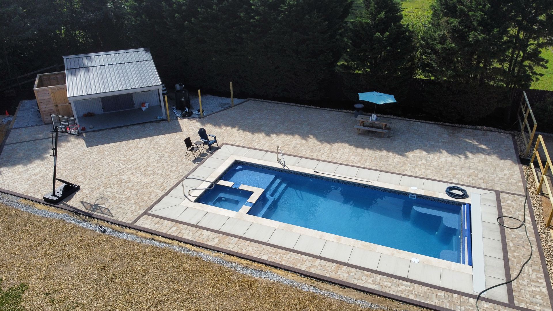 An aerial view of a backyard featuring a rectangular blue pool with a built-in spa, a shaded patio area, and a stone deck.