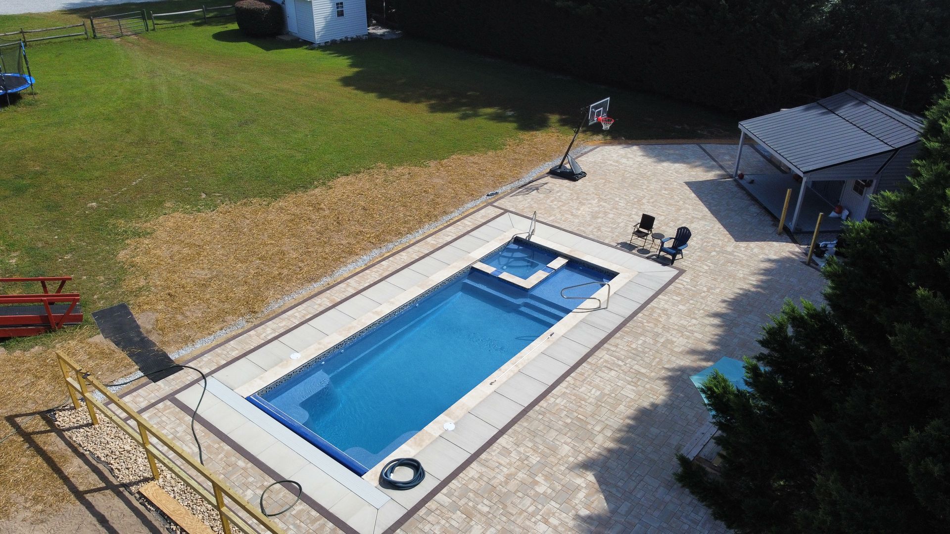 An aerial view of a rectangular blue swimming pool with a built-in spa, surrounded by a light-colored stone patio.