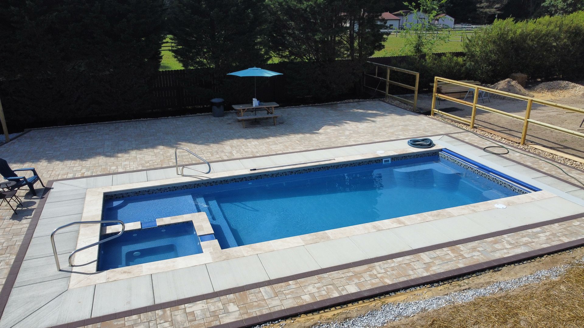 A rectangular blue swimming pool with an integrated spa, surrounded by a light-colored paved patio and a grassy yard.
