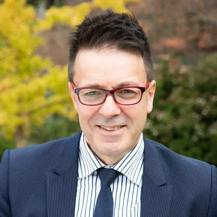 Man in red glasses and suit smiling outdoors with blurred autumn trees behind him