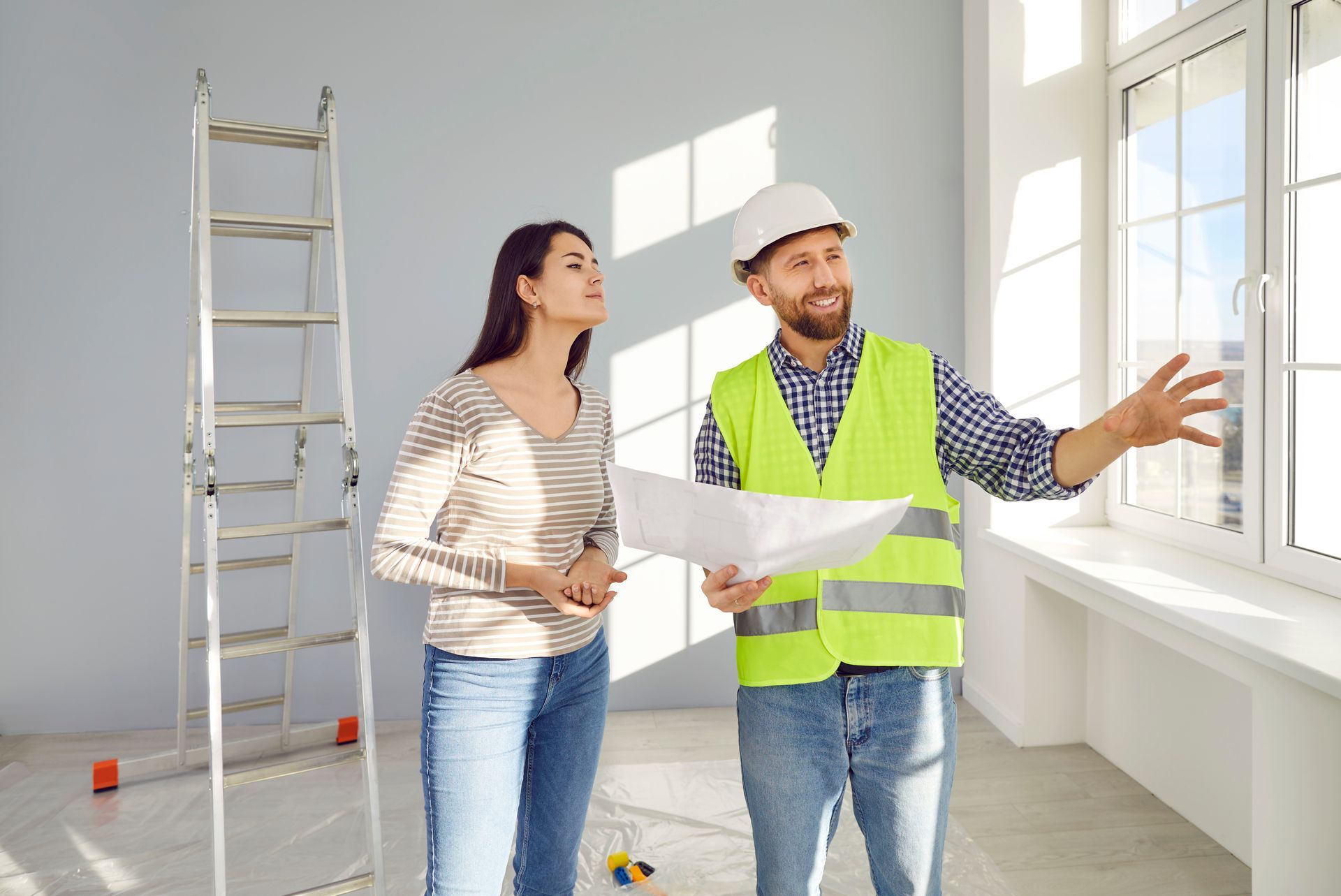 Two people reviewing renovation plans in a bright room with a ladder and window work area.