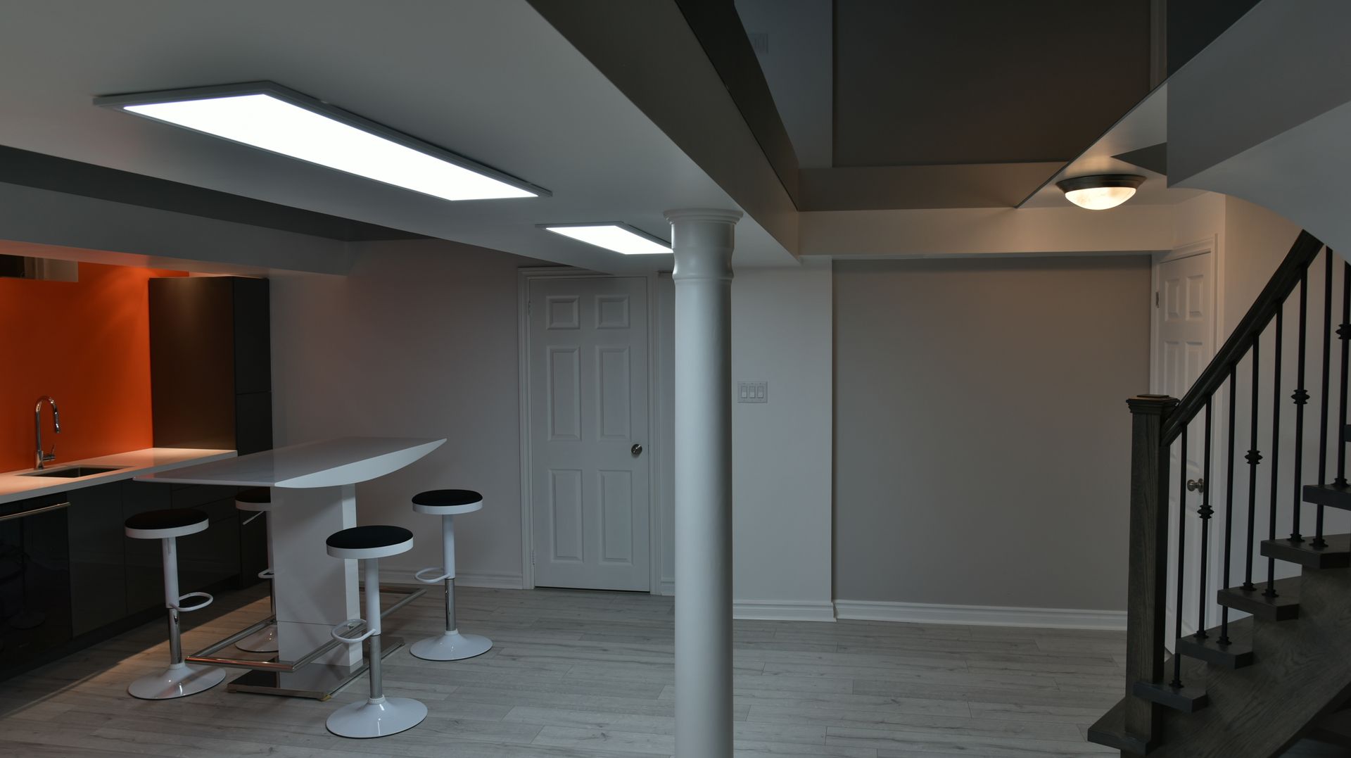 Modern open-concept basement with bar stools, staircase, white doors, and recessed lighting.