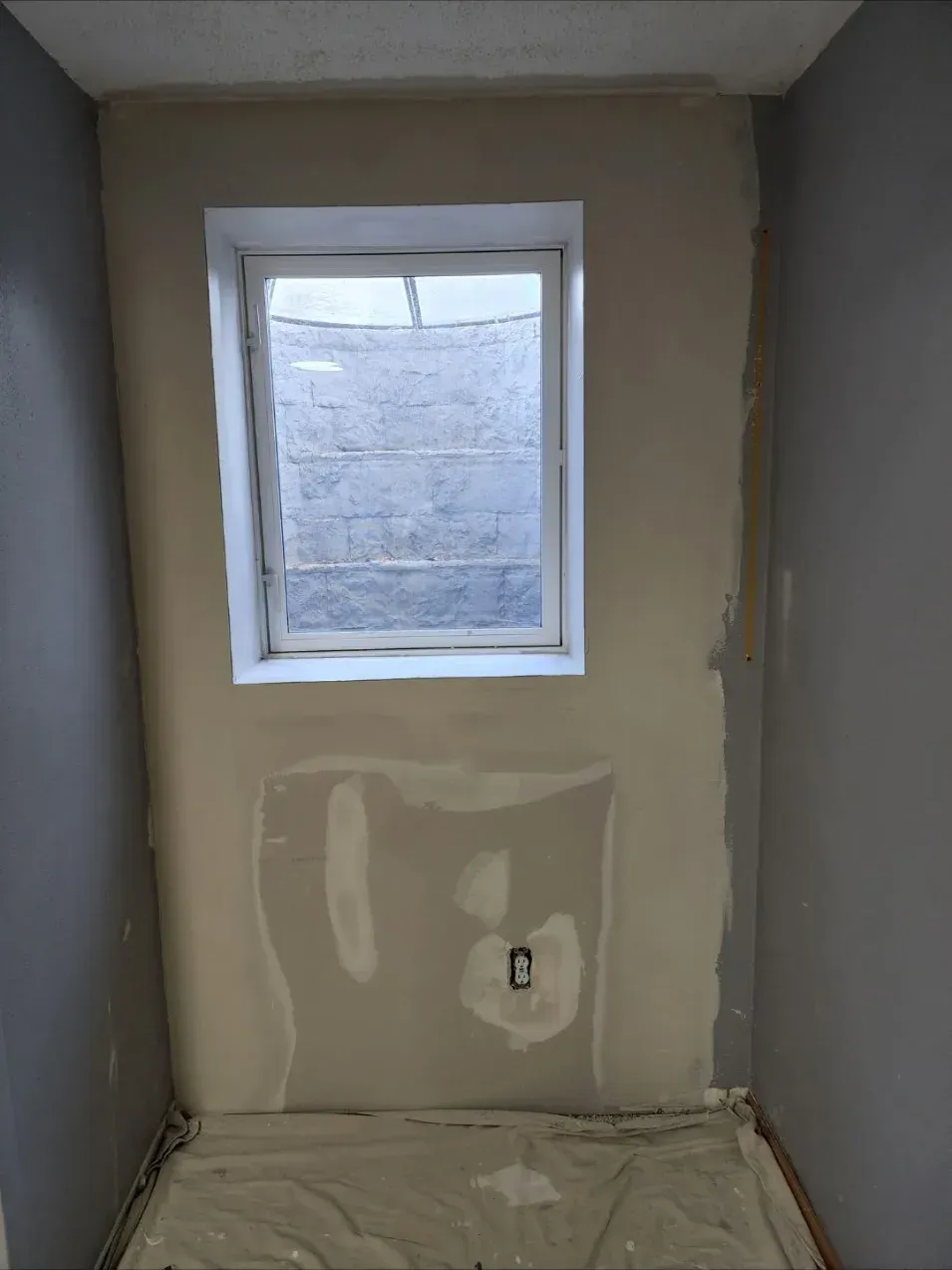Before repair: unfinished drywall patch with visible seams, tape lines, and an exposed outlet around a basement egress window in a Janesville, WI home