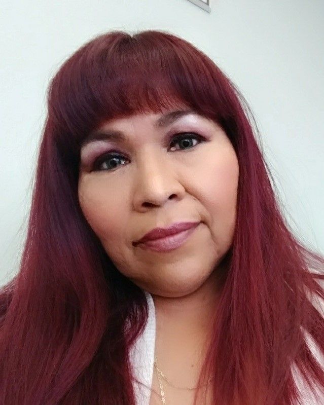 Close-up portrait of a person with burgundy hair and bangs, wearing a white top and necklace, against a light background