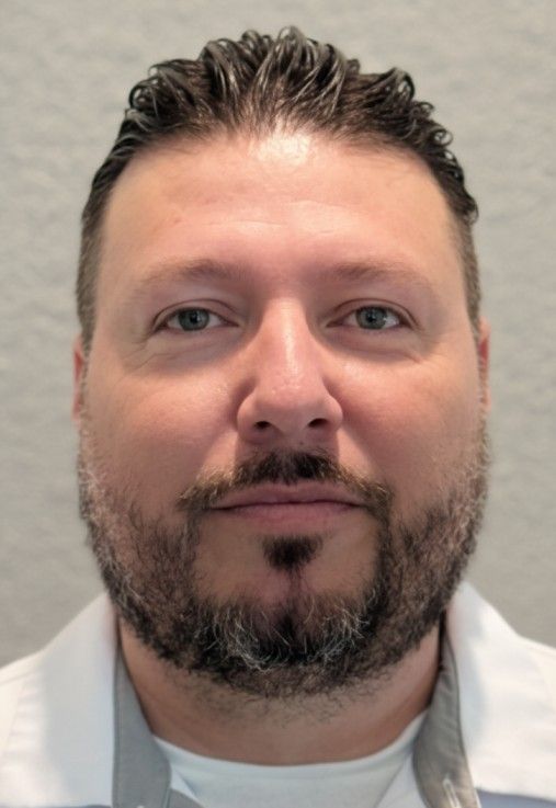 Portrait of a man with short dark hair and a beard, wearing a white shirt against a gray background