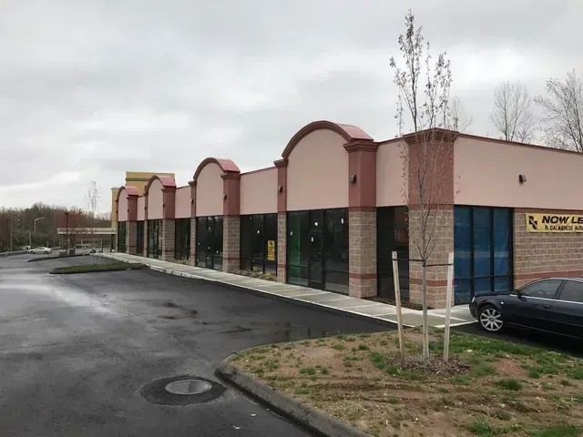 Empty storefronts in a strip mall on a rainy day, with a parked car and young trees.