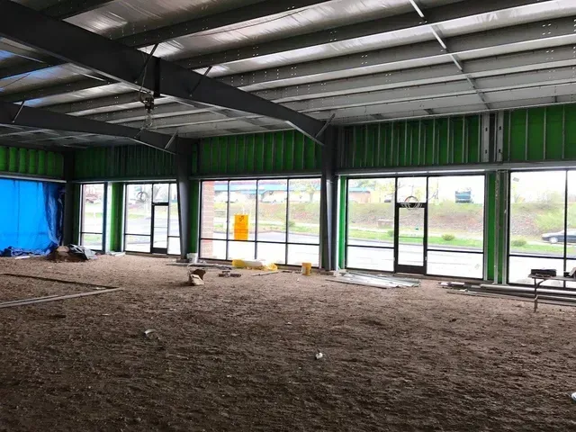 Empty industrial interior with green walls, large windows, and a dirt floor under exposed beams