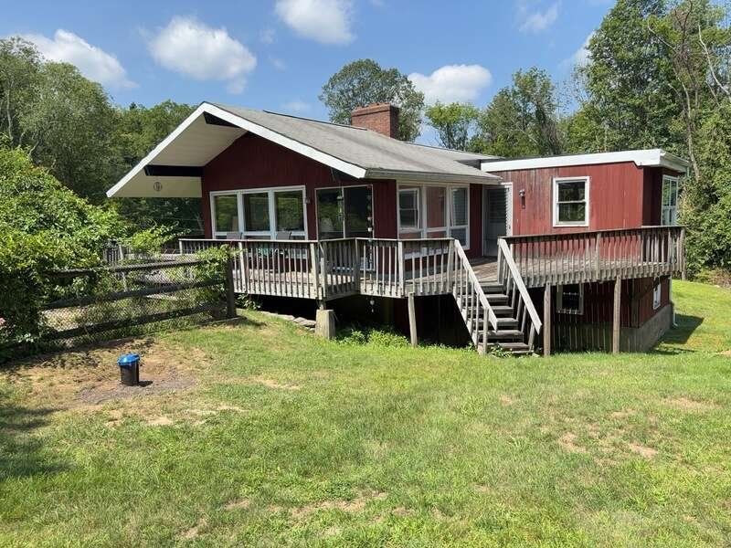Red house with a covered porch and stairs in a grassy yard, surrounded by trees under a blue sky