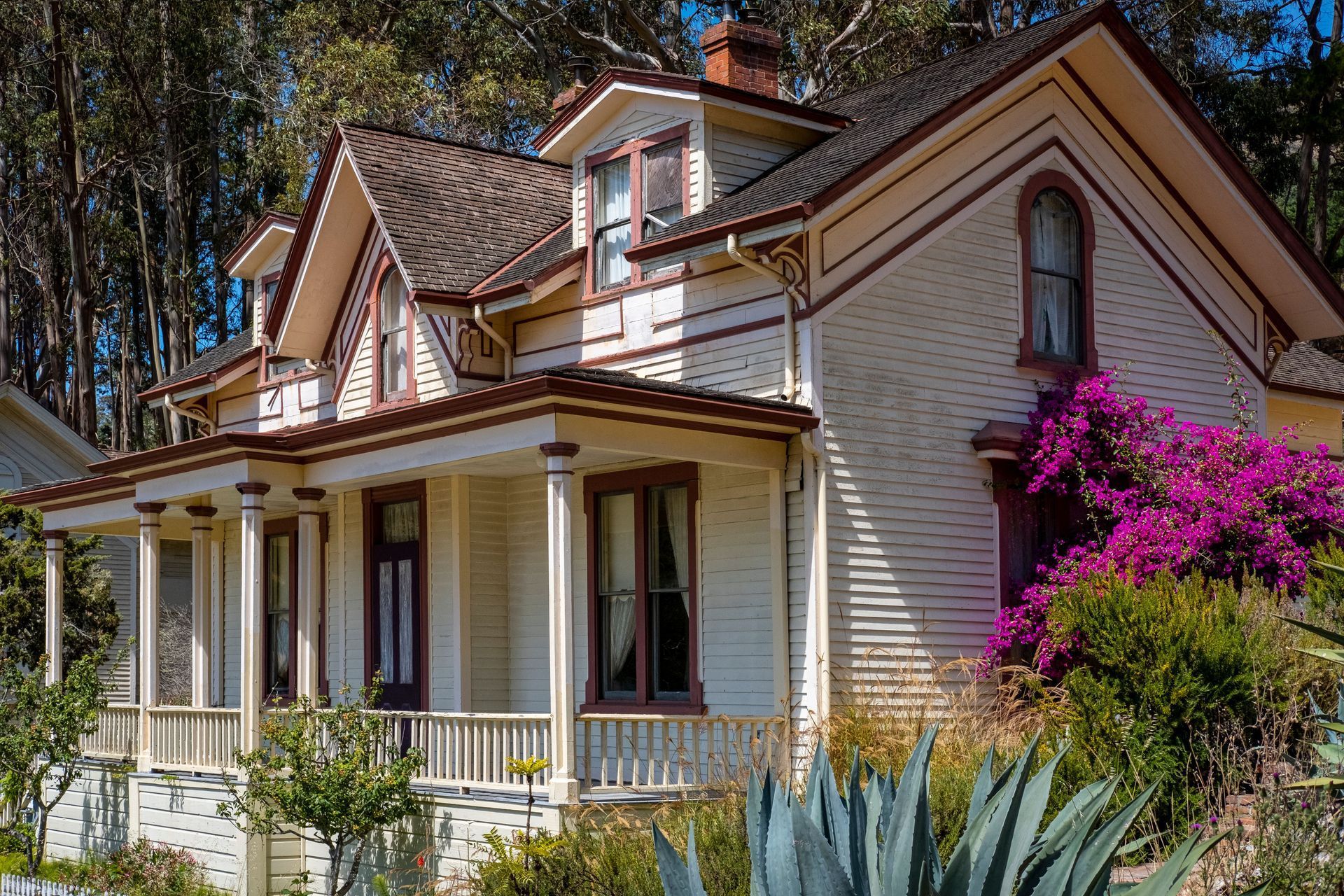 Victorian-style house with cream siding, porch, and bright purple bougainvillea in a wooded setting