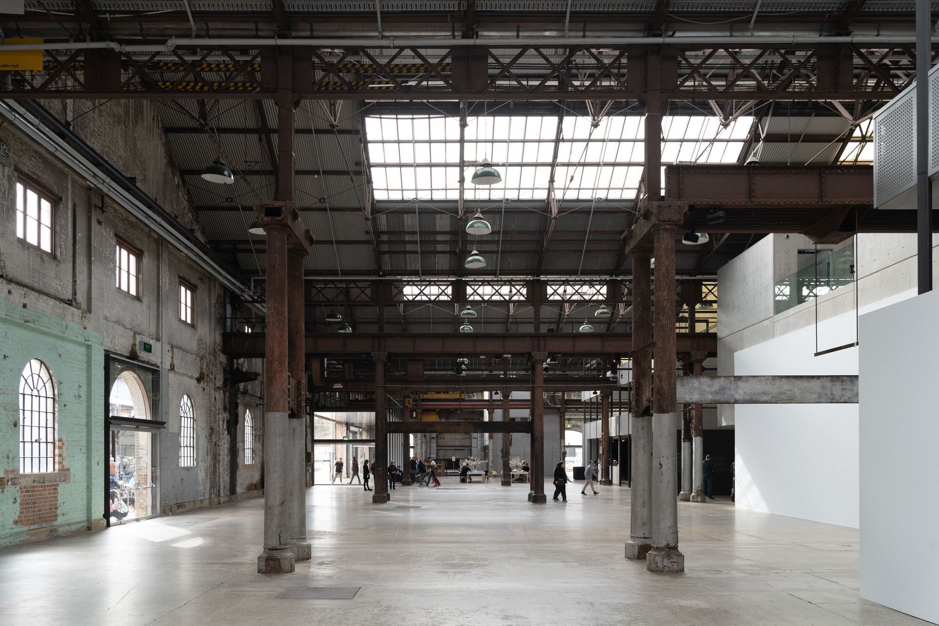 Empty industrial hall with exposed steel beams, large windows, and a few people in the distance