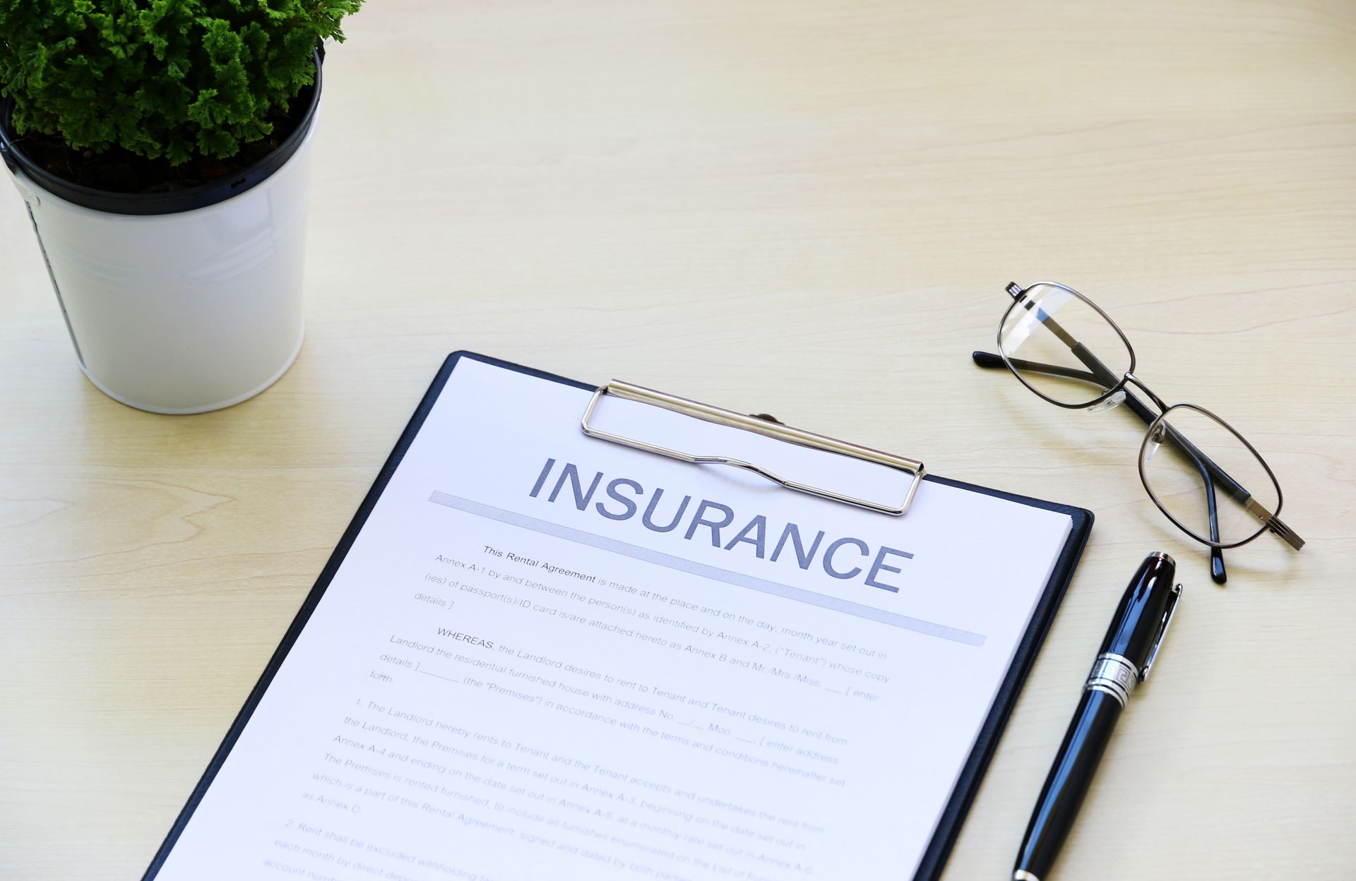 Insurance document on clipboard with pen and eyeglasses on a desk, beside a small potted plant