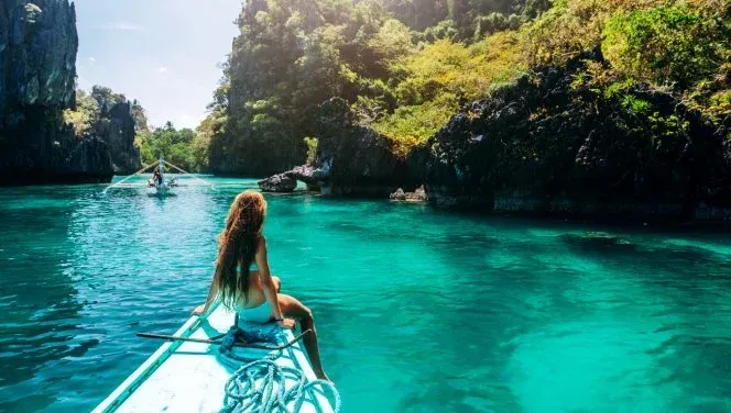 A woman gazes upon a blue-green lagoon. The warm sun hits the lush green rocky sides of the bank. 