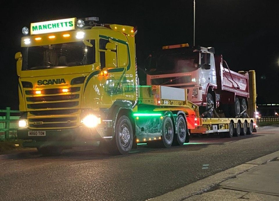 Manchetts Yellow Scania truck towing a black-and-white bus on a tree-lined street