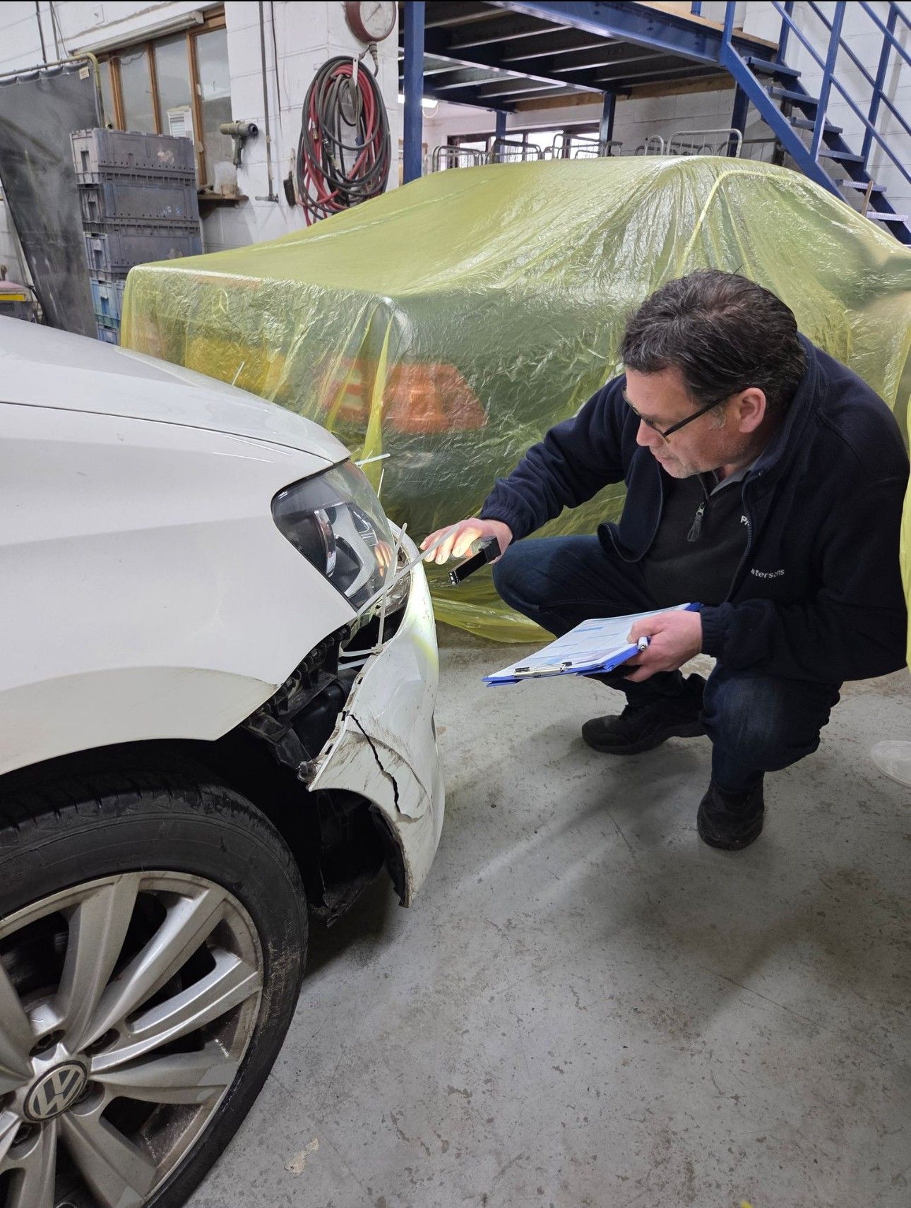 Mechanic inspecting damage on a white car’s front bumper in a workshop, holding a clipboard.