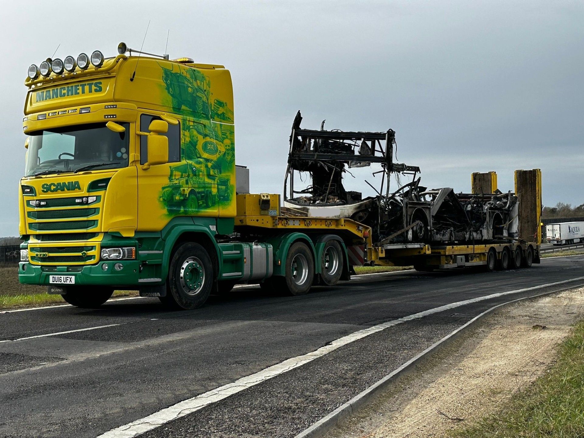 Yellow and green Manchetts semi truck hauling a charred vehicle frame on a flatbed trailer
