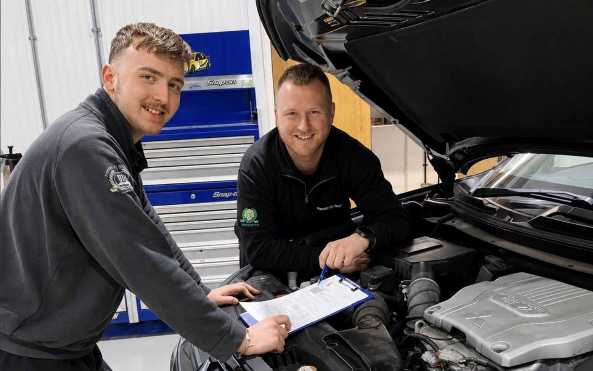 Two mechanics smiling beside an open car hood, checking a clipboard in a garage