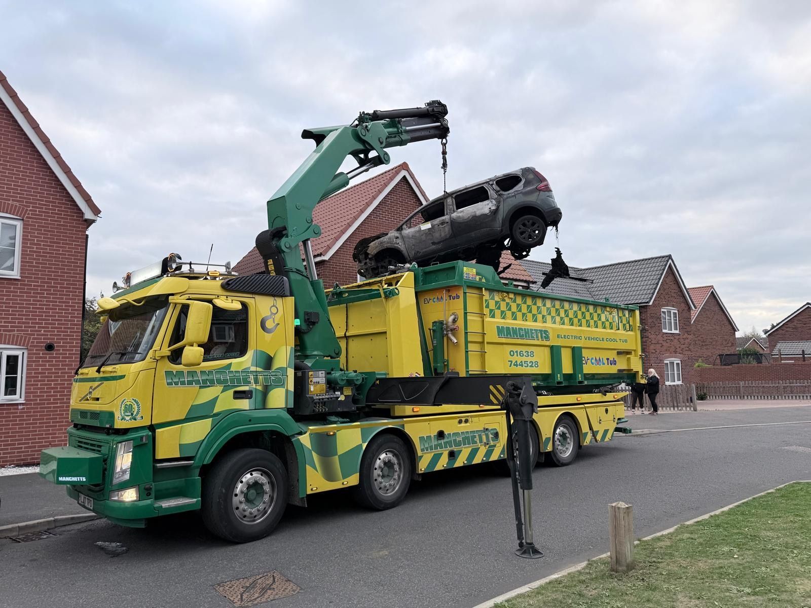 Manchetts Tow truck lifting a black car onto a green and yellow recovery truck on a residential street