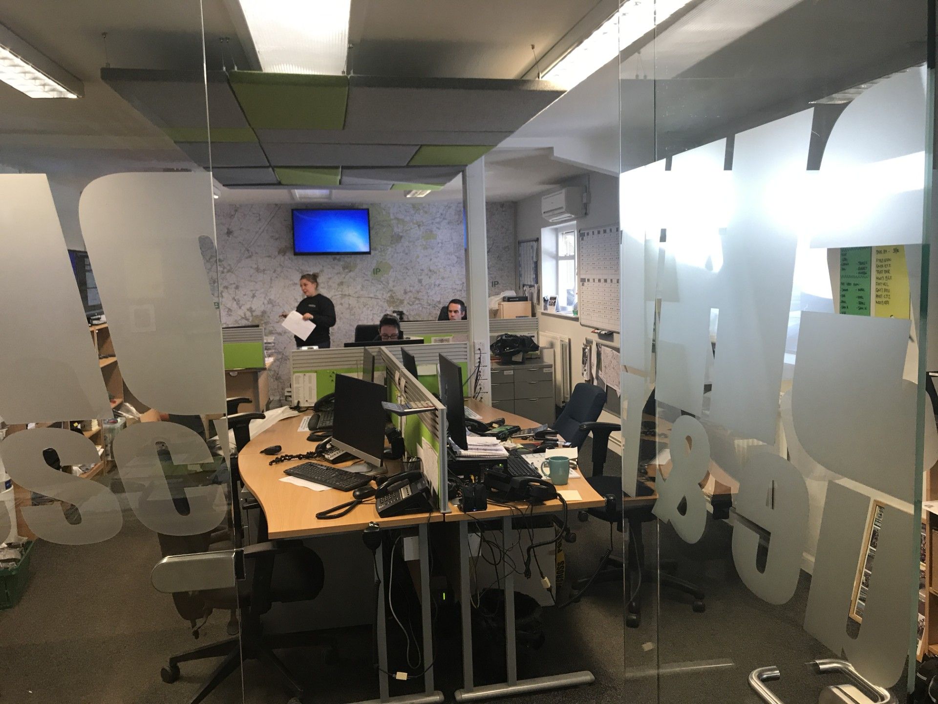 Open office with desks, computer monitors, and two people near a wall-mounted TV.