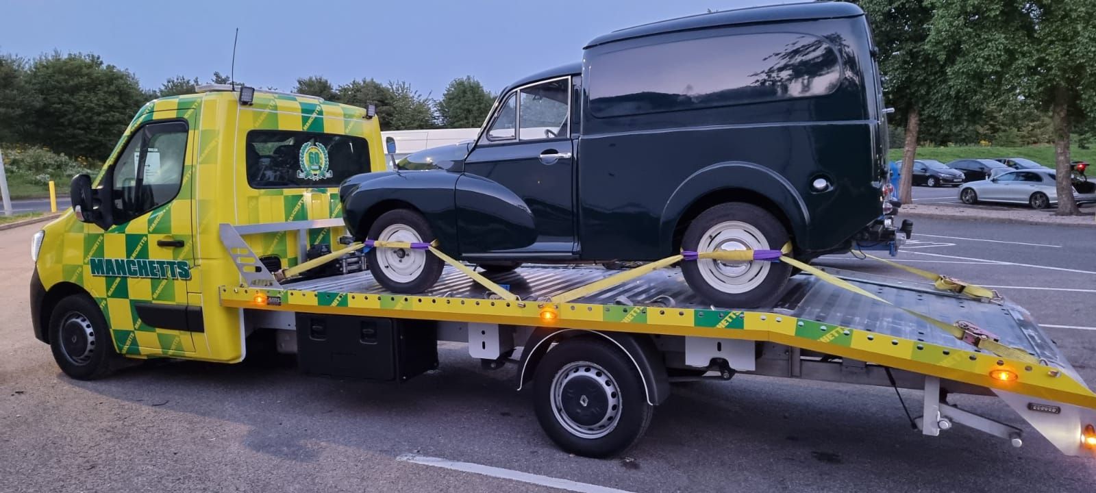 Tow truck hauling a black vintage van on a yellow flatbed in a parking lot
