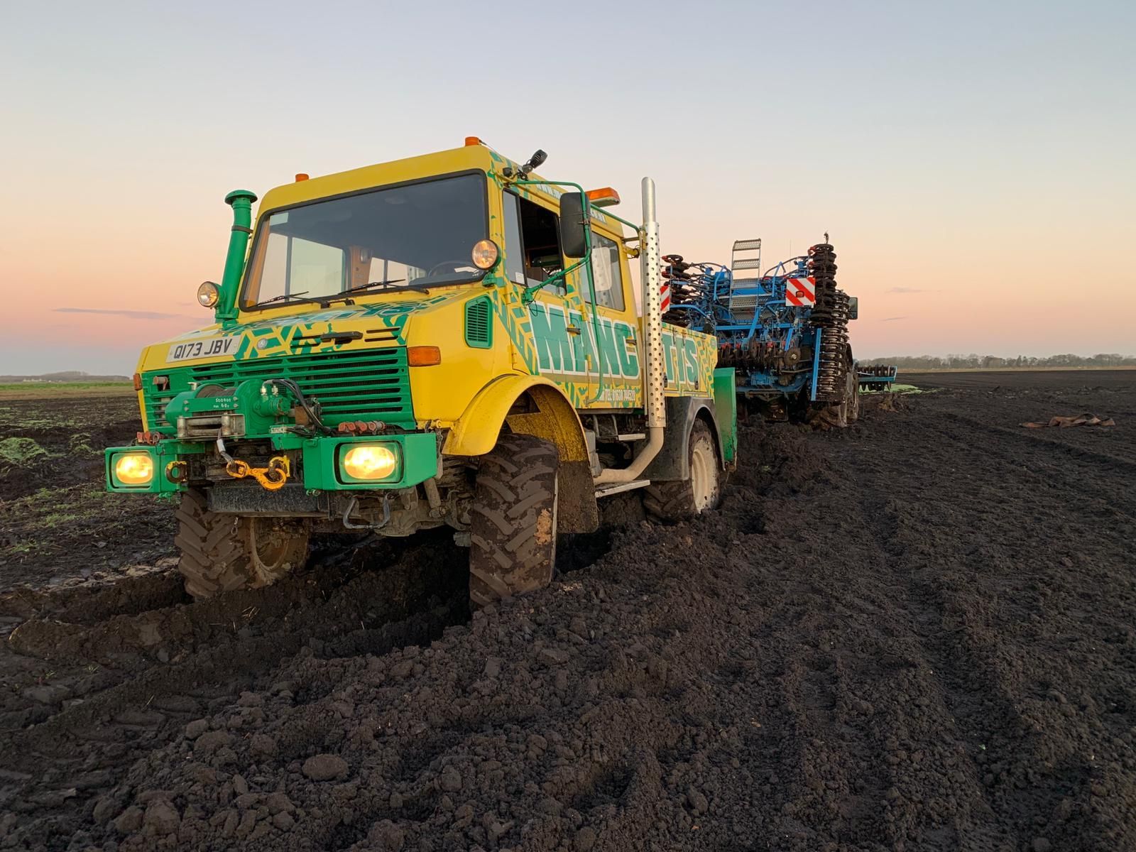 Yellow and green Manchetts farm truck hauling equipment along a muddy field road at sunset