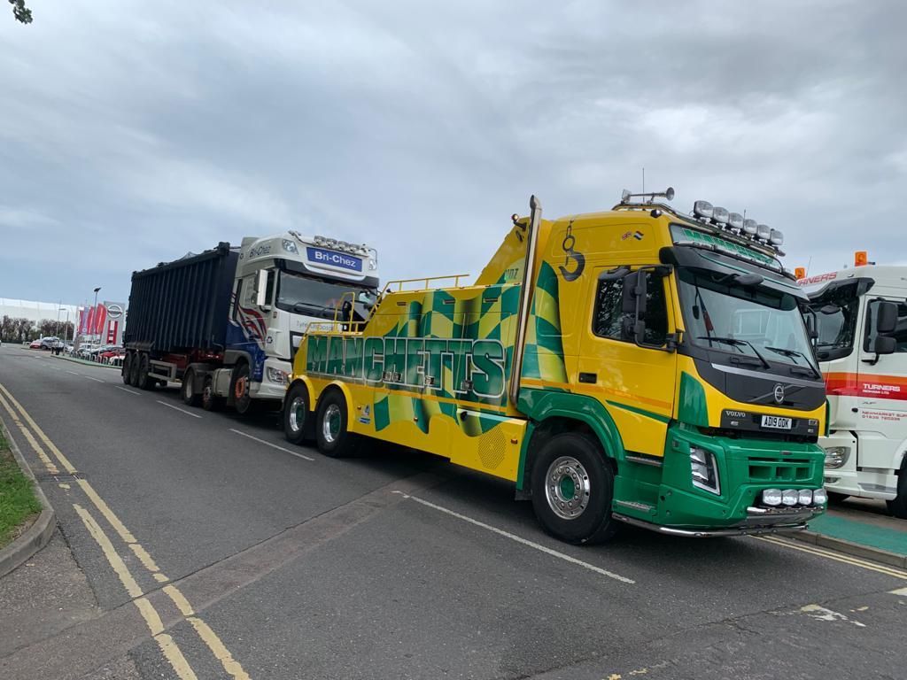 Bright green and yellow Manchetts tow truck hauling a black trailer on a roadside under cloudy skies