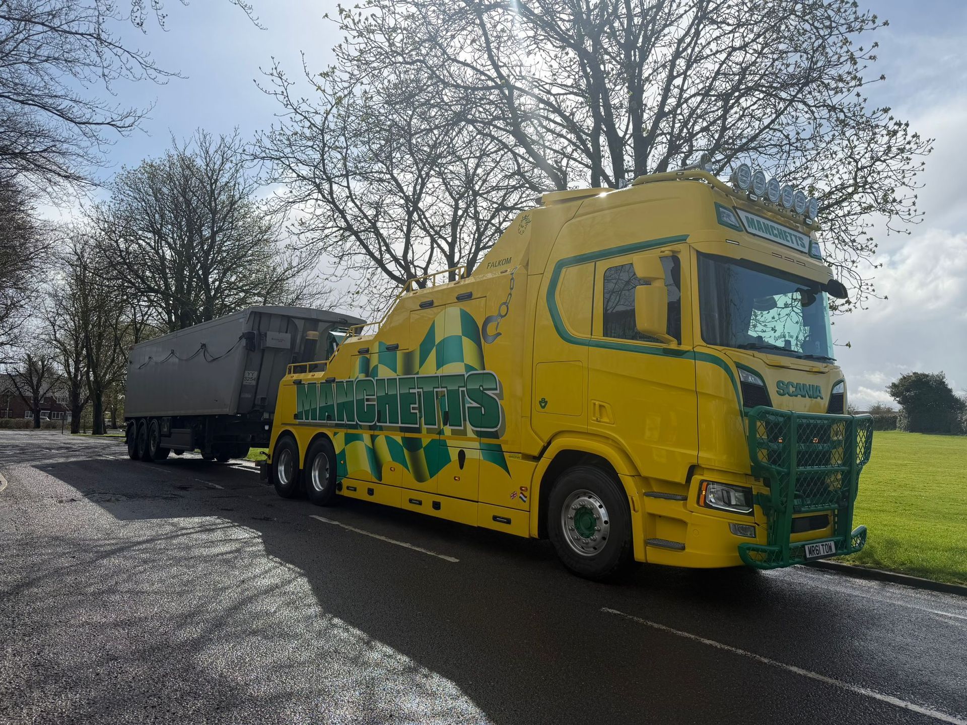 Yellow Manchetts decorated truck towing a dark trailer on a road beside trees under a cloudy sky