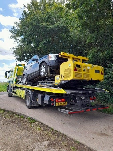Tow truck carrying a black car and yellow equipment on a roadside near trees