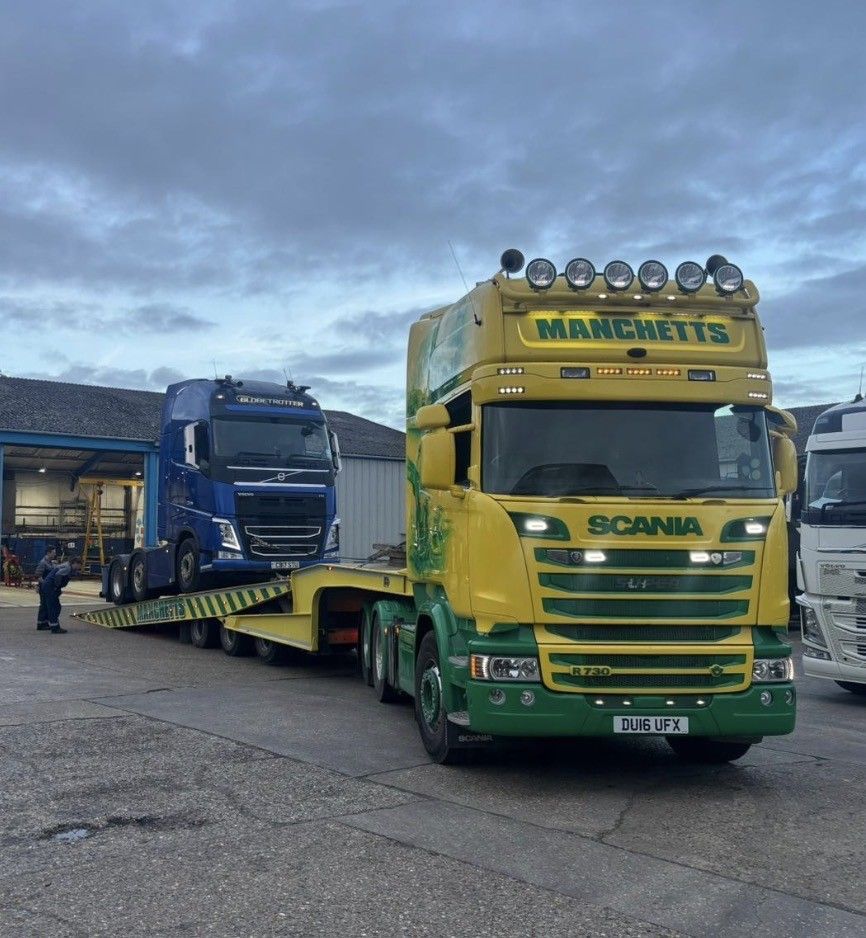 Yellow and green Manchetts Scania tow truck hauling a blue truck in a yard under a cloudy sky