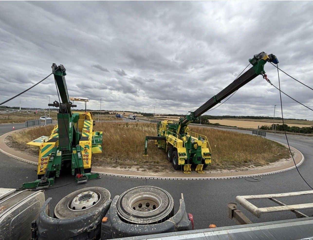 Two green Manchetts crane trucks lifting large tires at a roundabout construction site under cloudy skies