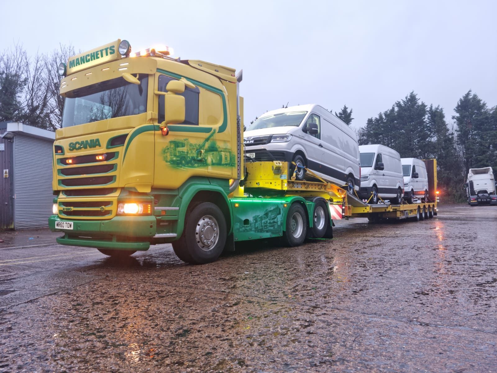 Yellow-green Manchetts tow truck hauling a white van on a wet road in rainy weather