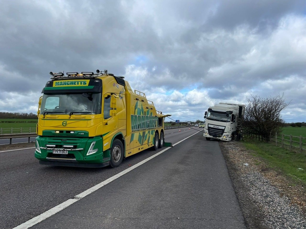 Yellow-green Manchetts tow truck on a rural road beside a tilted white truck under cloudy skies