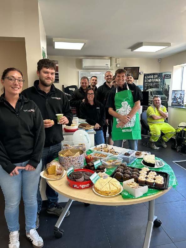 Group of people behind a table of desserts and baked goods in a bright indoor room.
