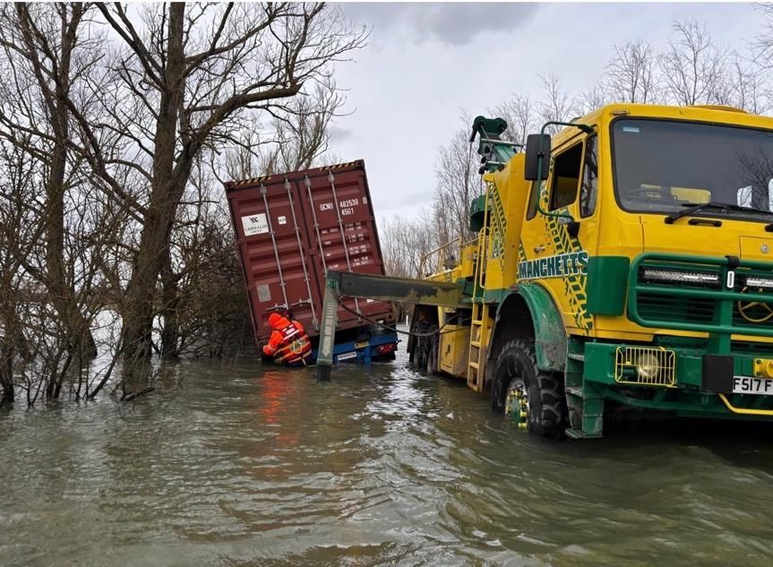 Manchetts Tow truck in floodwater lifting a red cargo container beside bare trees