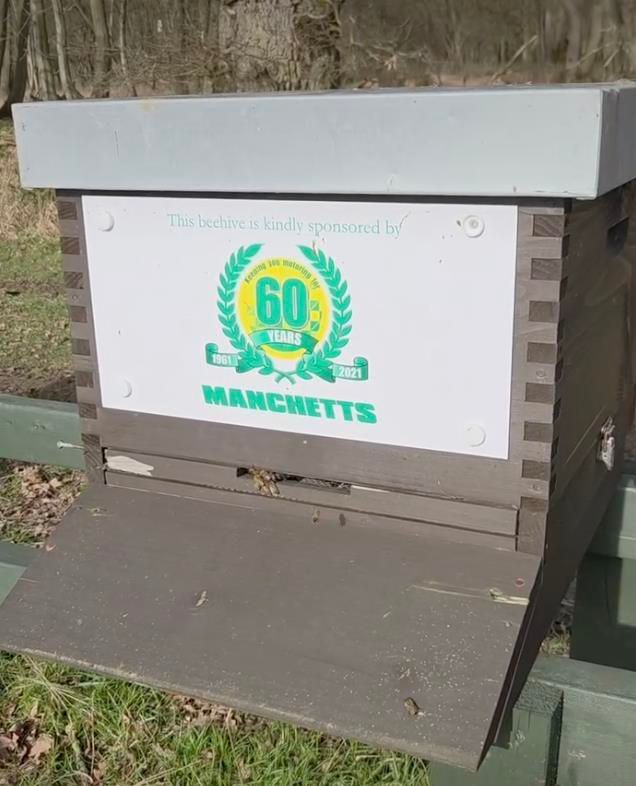 Wooden beehive box with a “60 years” Manchetts sign in a wooded outdoor area