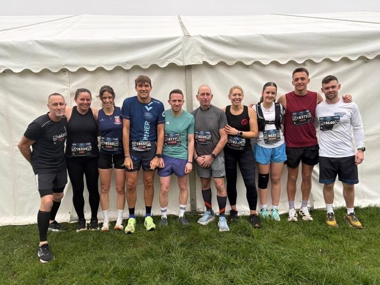 Group of runners posing in front of a white event tent on grass, wearing race shirts and medals.