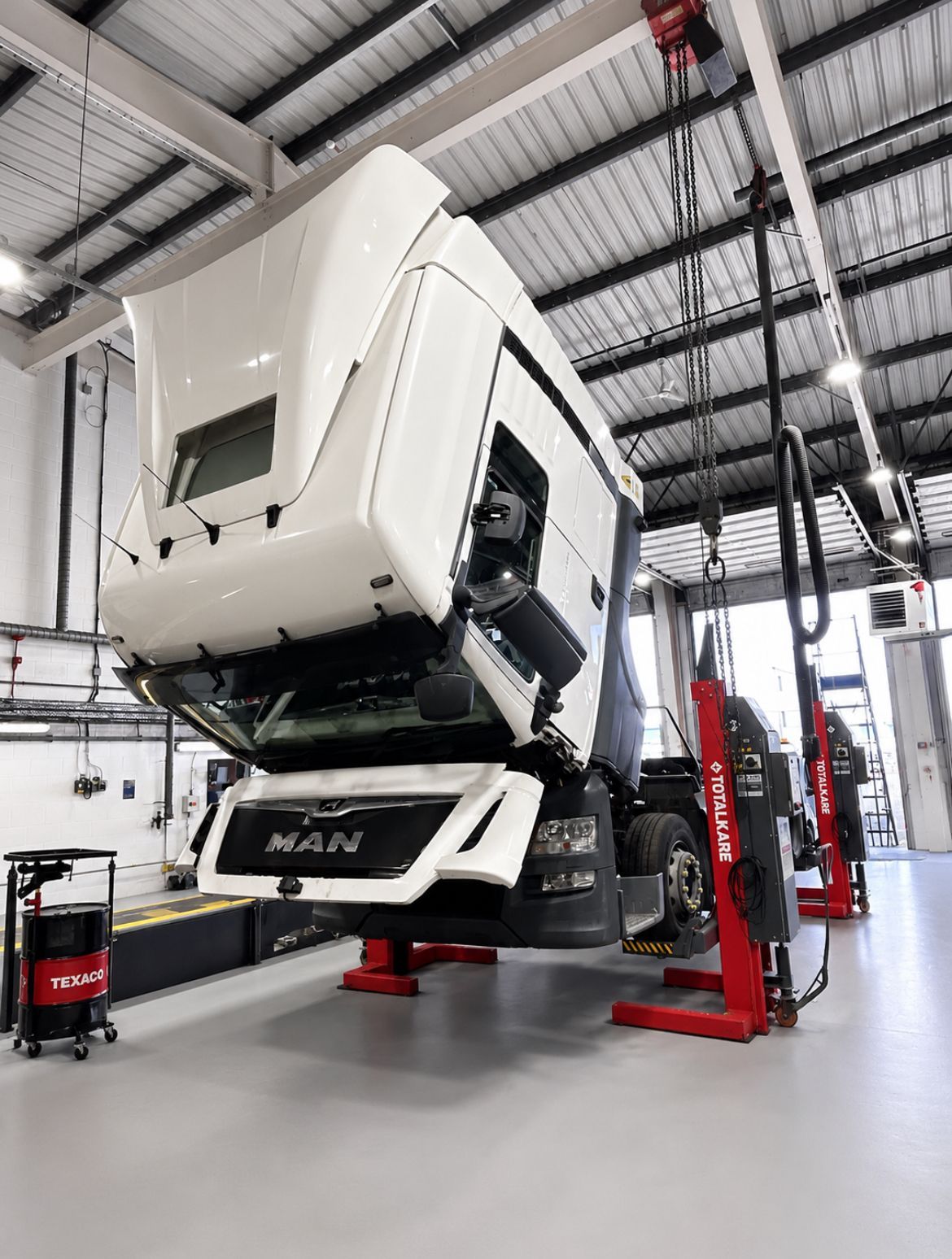 White semi-truck cab elevated on red hydraulic lifts inside a bright automotive workshop.