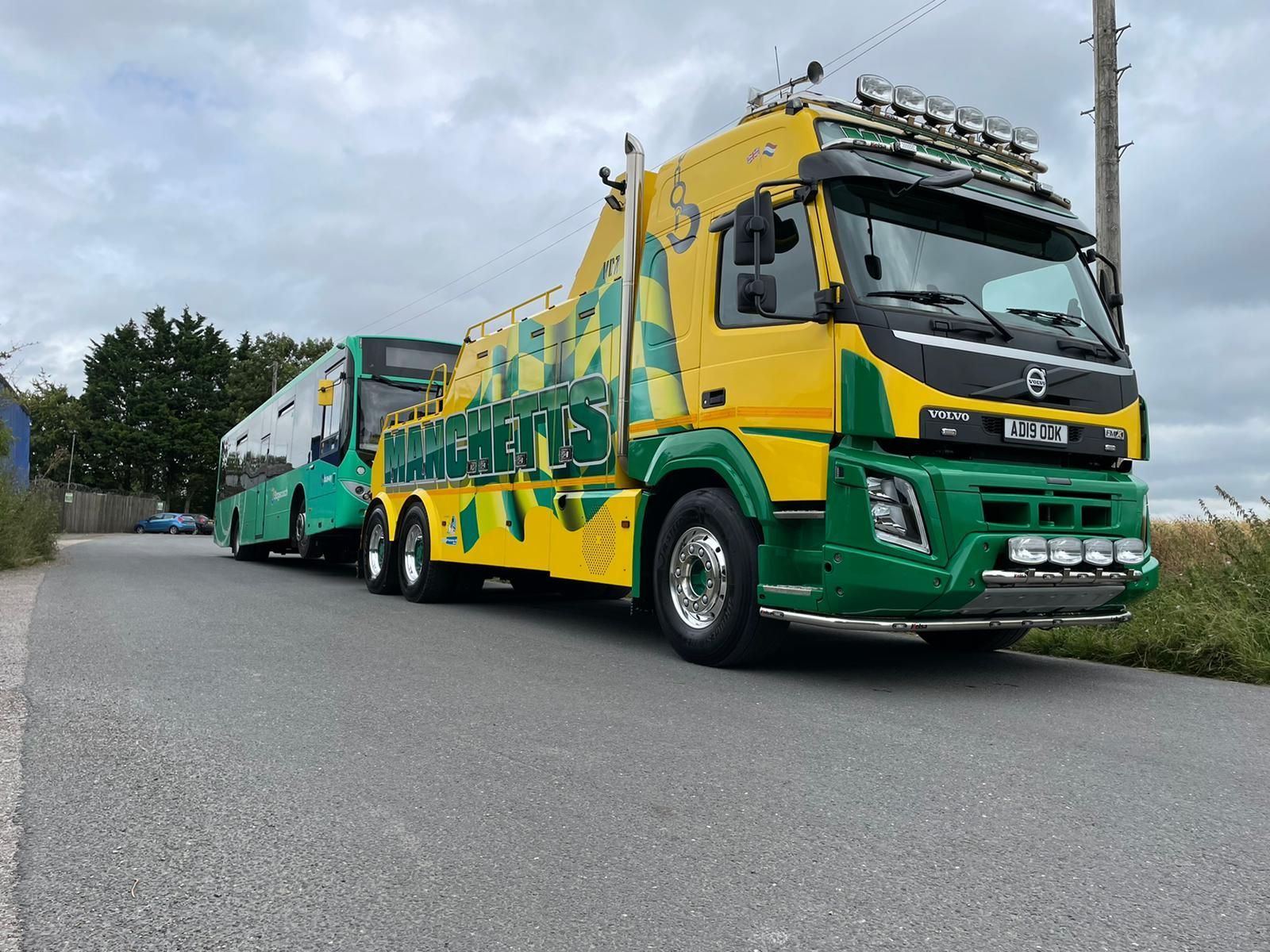 Green and yellow Manchetts semi-trucks lined up on a road, viewed from the front-left side.