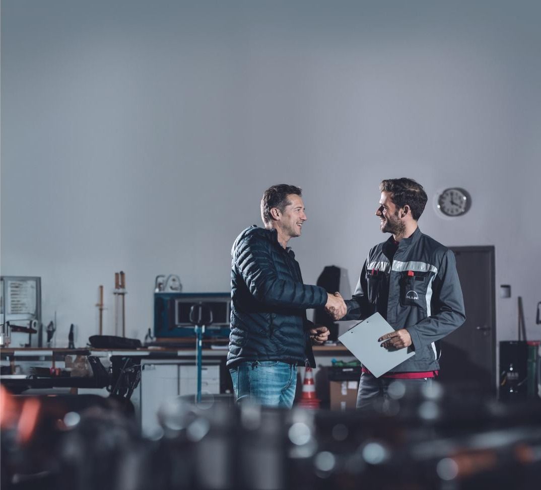 Two people shaking hands in a workshop, one holding a clipboard, with machinery blurred in the background.