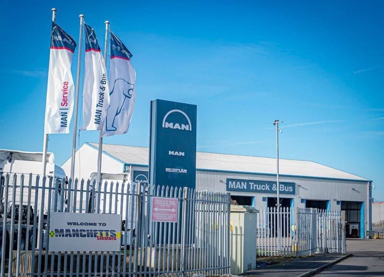 Manchetts Truck dealership lot with MAN signs, white flags, and a warehouse building under a blue sky