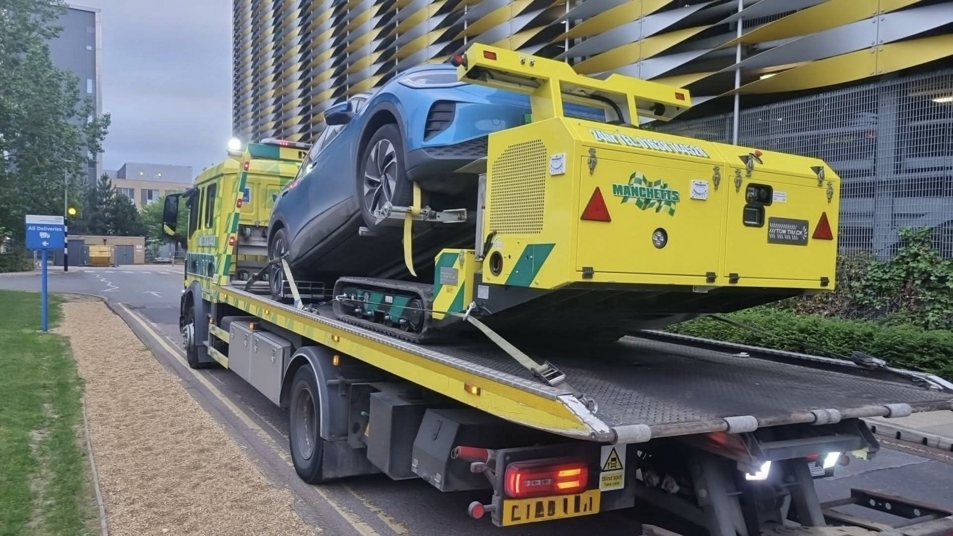 Manchetts Tow truck carrying a blue sports car on a city street beside modern buildings