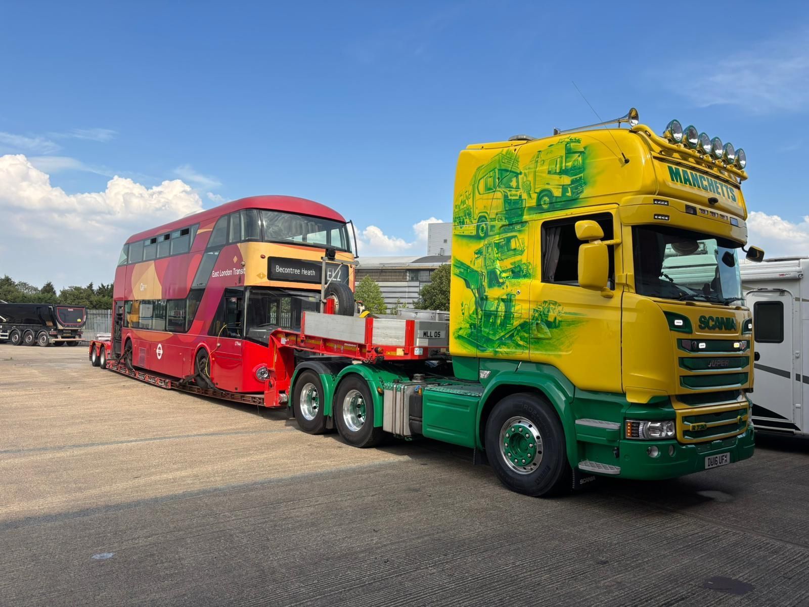 Yellow and green semi truck carrying a red double-decker bus in an open lot under a blue sky
