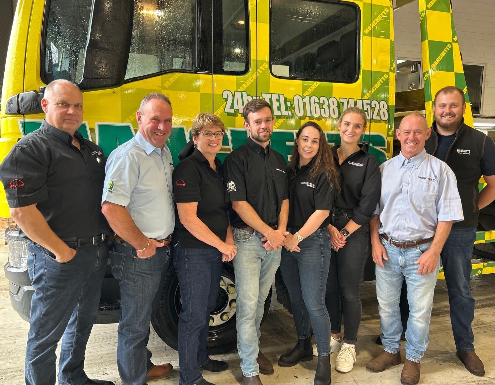 Group of coworkers standing in front of a yellow-green Manchetts truck in a garage, posing for a photo