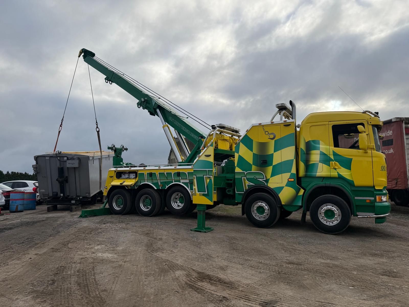 Green-and-yellow tow truck lifting a gray container in a gravel lot under cloudy skies