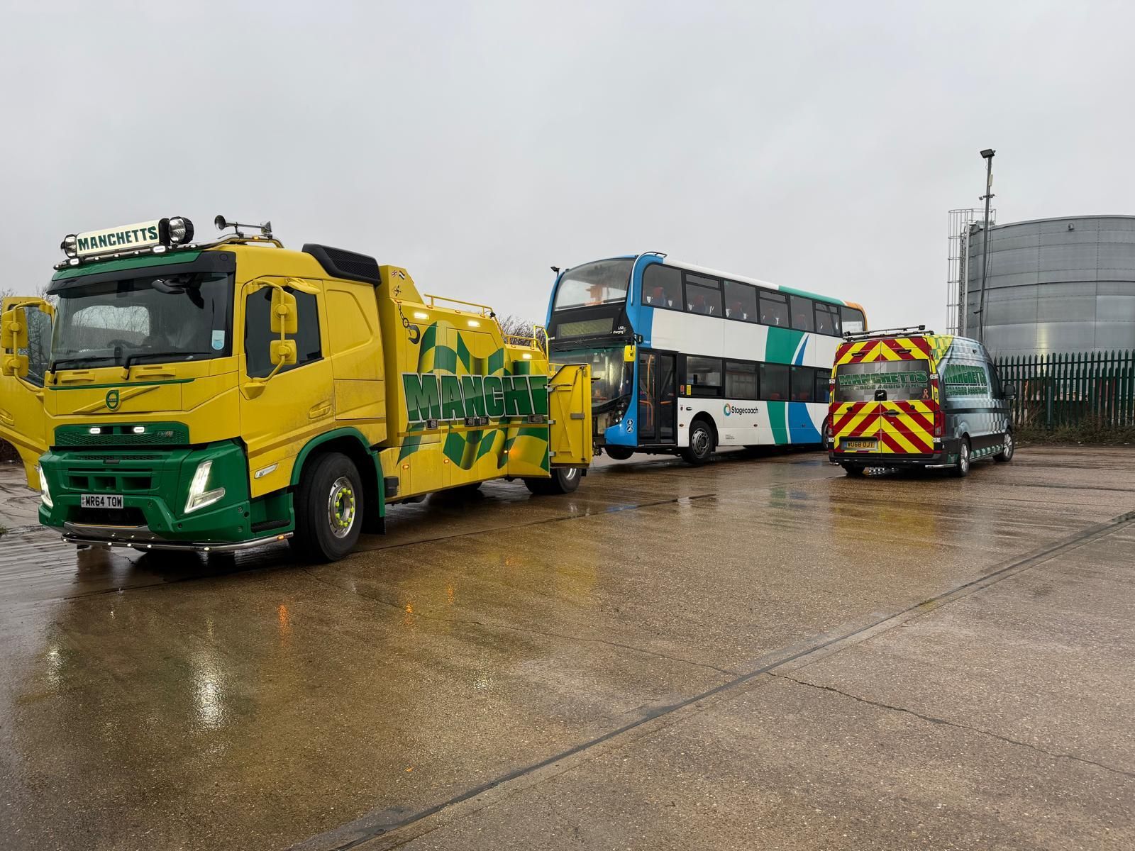 Manchetts Yellow and green tow truck with a double-decker bus on a wet lot, industrial tanks in background