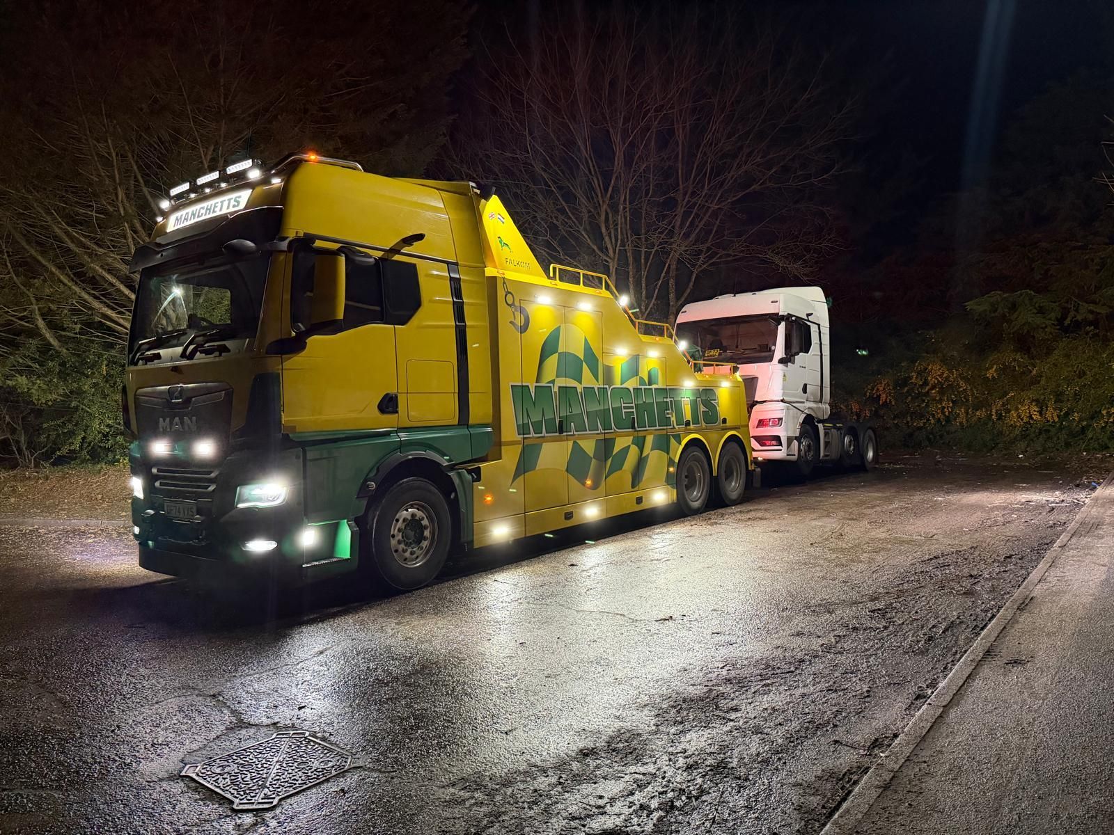 Manchetts Yellow tow truck hauling a white truck on a wet road at night