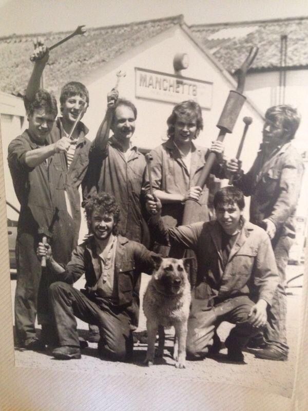 Group of people posing outdoors with tools and a dog in front of a building.