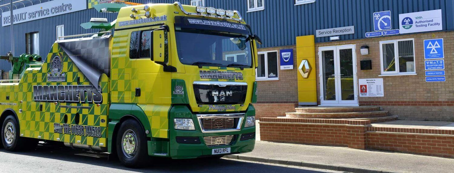 Green Manchetts tow truck parked outside a building, with blue and yellow service signage in the background