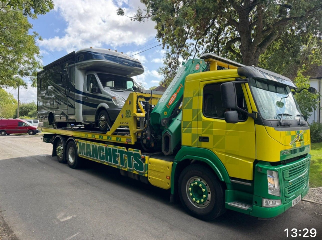 Green and yellow Manchetts tow truck carrying a black RV on a tree-lined street