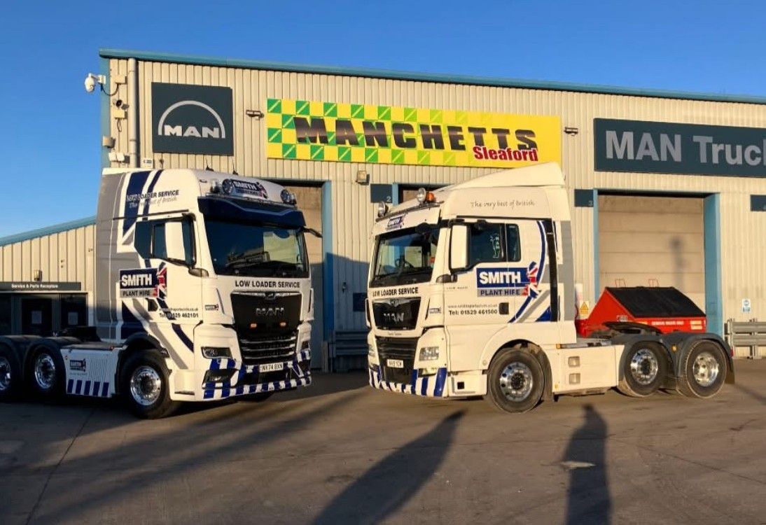 Two white MAN trucks parked outside a Manchetts truck dealership with a yellow sign and blue sky