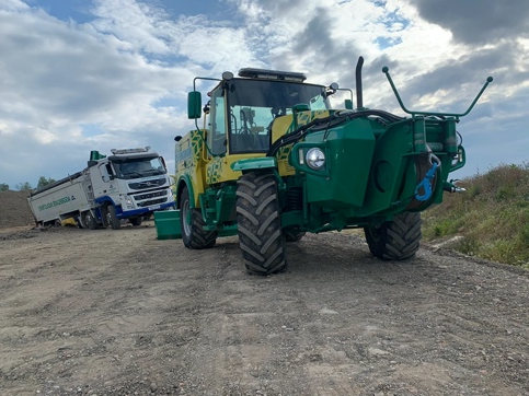 Green Manchetts tractor on a dirt road with a blue dump truck in the background under a cloudy sky